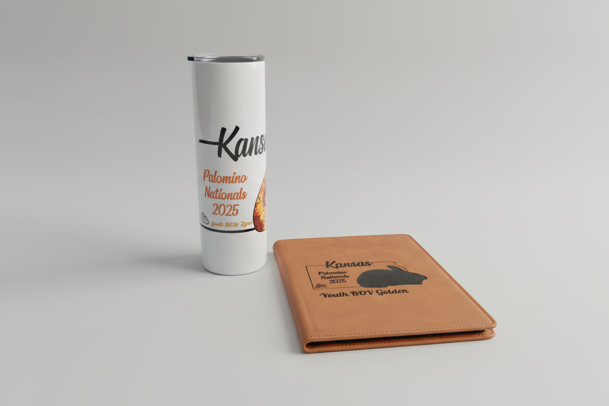 White tumbler with 'Kansas Palomino Nationals 2025' text and a brown notebook with similar text on a metallic surface.