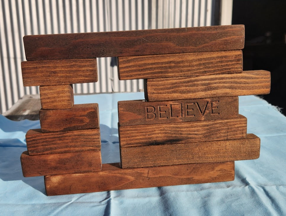 Wooden sculpture with 'BELIEVE' engraved on it, placed on a blue surface.