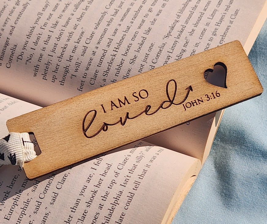 Wooden bookmark with 'I AM SO LOVED' text and heart cutout on an open book.